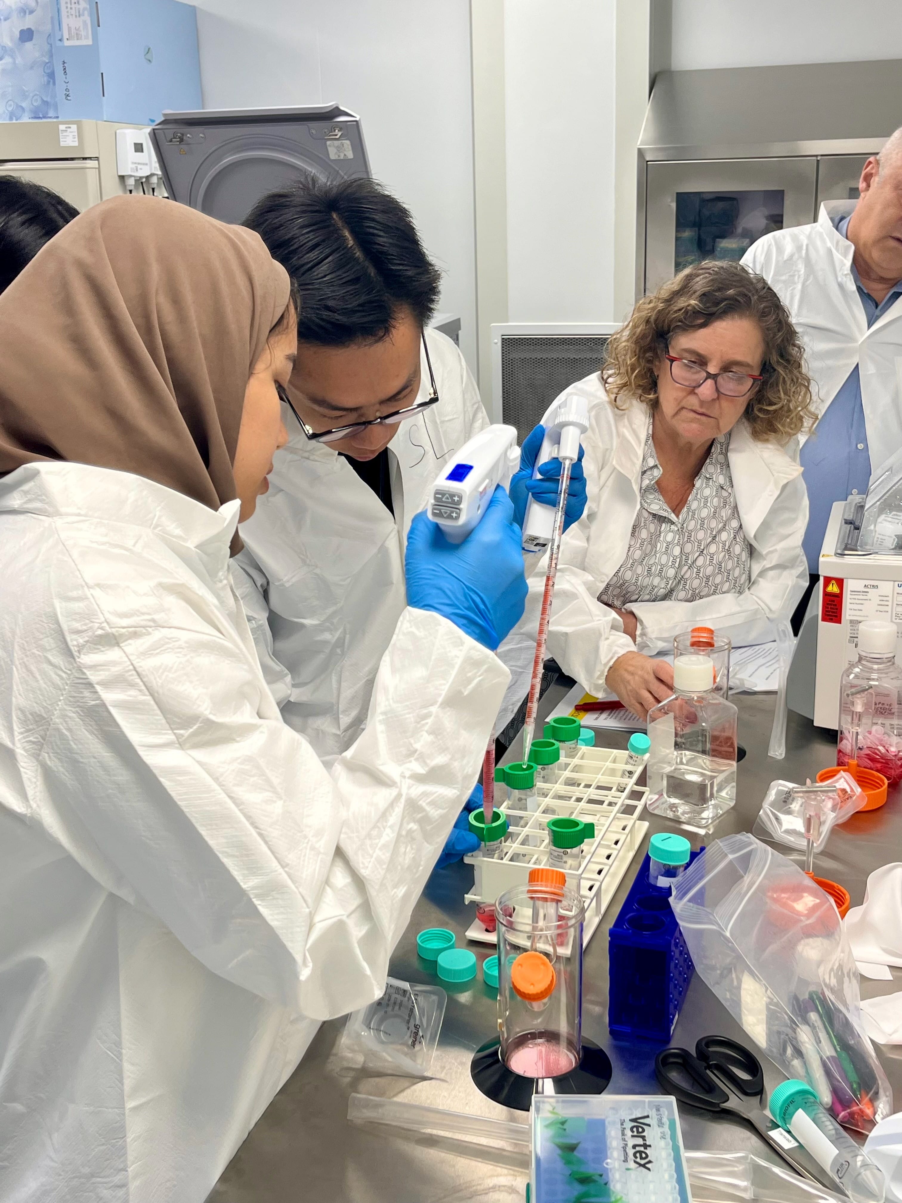 Participants in laboratory protective attire pipetting solutions in a hands-on cell culture training session, under the guidance of facilitators in a research laboratory.